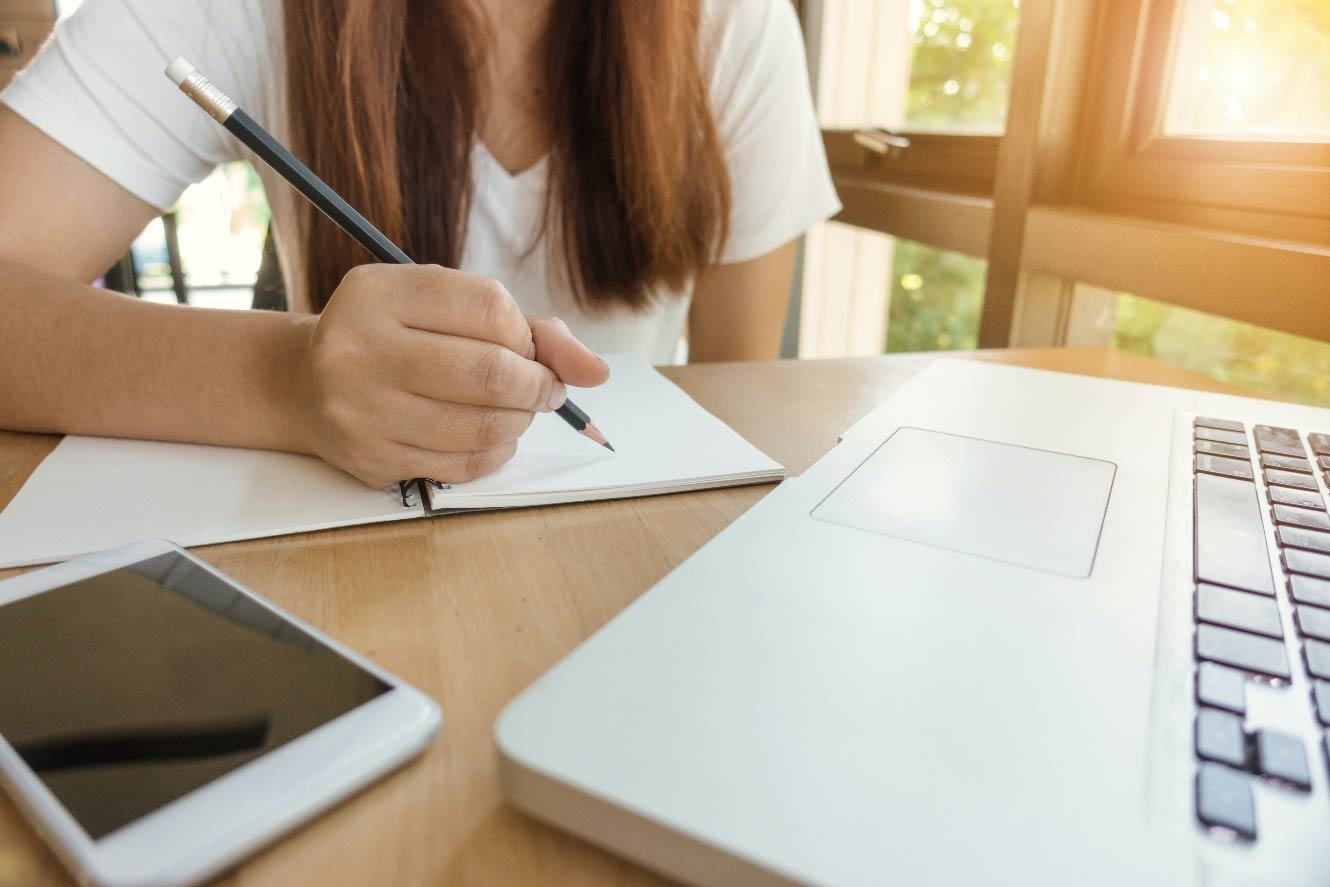 girl writing on blank notebook with laptop and mobile phone on the desk 